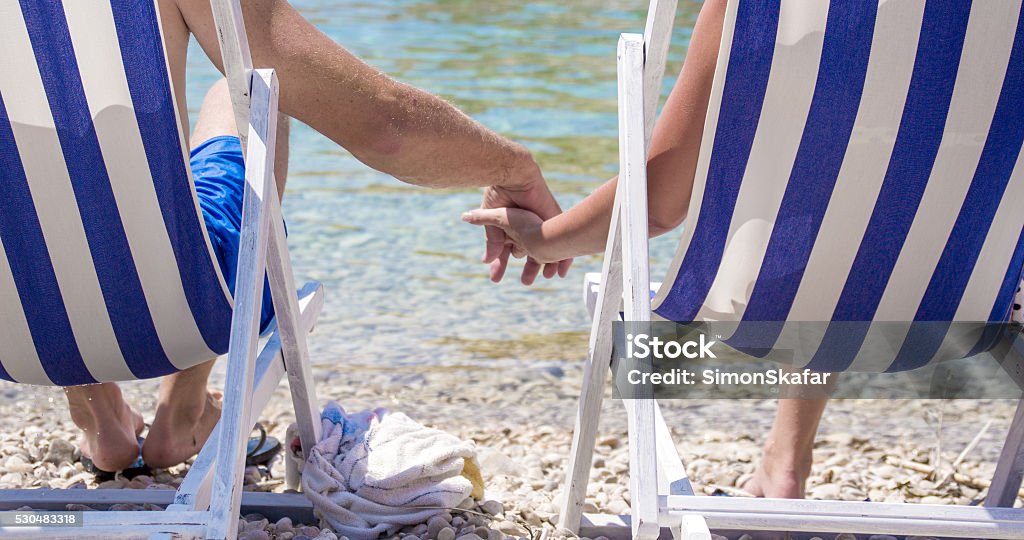 Couple holding hands on deck chairs at Vis island Low section of couple holding hands while relaxing on deck chairs at beach. Male and female tourists are visiting Vis Island during vacation. They are on shore at island. Blue Stock Photo