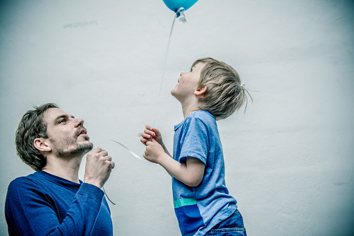 Happy father and son of four together indoors, Paris, France. They are both in blue shirts and jeans. The father is crouching, they are both holding a string with a blue ballon. The boy is looking at the baloon, his father is looking at him. Copy space. Nikon D800, full frame, XXXL. iStockaLypse Paris 2016.