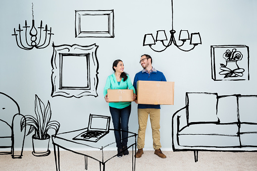 Mid adult Hispanic couple imagine their house as they stand in an empty living room. Digital artwork imposed on the photo includes picture frames, sofa, chair, coffee table with laptop and lighting equipment. The couple hold boxes and are looking at one another as they daydream. Custom artwork copyright is wholly owned by Steve Debenport Imagery, Inc.