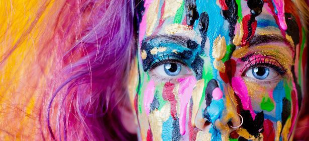 A close up photo of a woman with purple hair making direct eye contact with the camera while different colours of paint drip down her face. She has very striking, blue eyes and a nose ring.