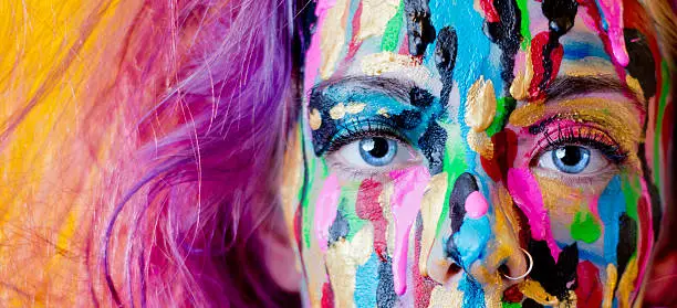 A close up photo of a woman with purple hair making direct eye contact with the camera while different colours of paint drip down her face. She has very striking, blue eyes and a nose ring.