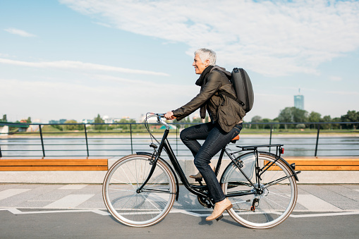 Mature businesswoman riding bicycle to and from her office. She is riding near river. Casual clothing with modern business backpack on her back. Riding city style bike.