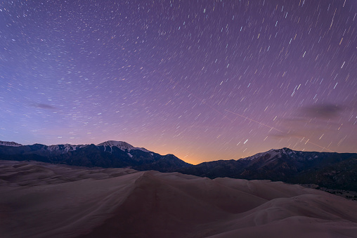 Star trails of spring night sky over snow peaks and great sand dunes at Great Sand Dunes National Park & Preserve, Colorado, USA.