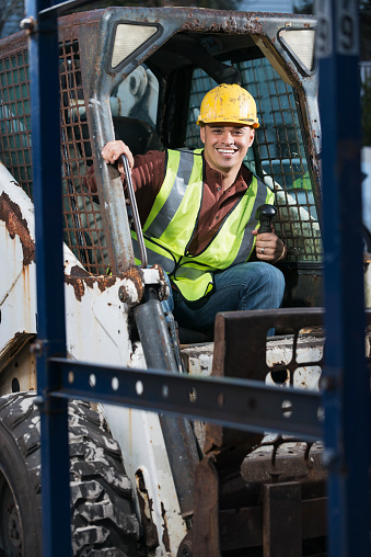 An Hispanic construction worker at a job site, sitting in a backhoe, wearing a yellow hardhat and safety vest. He is getting ready to climb out, smiling and looking at the camera.