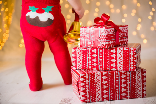 Composition of nicely ribbon wrapped and piled Christmas gift boxes in red and white paper motiv and baby half waist from the back in funny festive red tights with knitted Christmas muffin holding brass Santa bell in his right hand. Shot on Canon EOS 6D, 70mm, f2.8, ISO 250