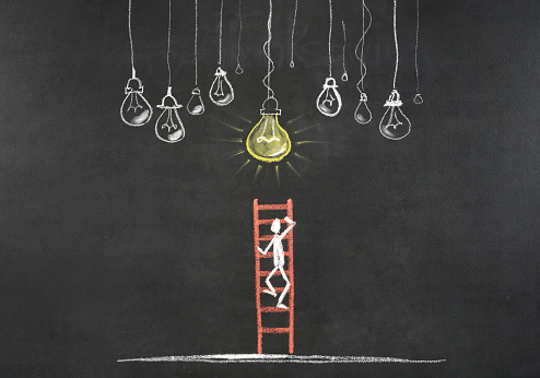 The chalkboard drawing shows light bulbs in a row with one glowing. Directly below a flat field where a ladder is stacked up towards the light bulbs. A man is climbing a ladder. The man is outlined in white, the ladder is red. The small light bulbs are outlined in white,the big light bulb is yellow. The glowing light bulb is a symbol of success.