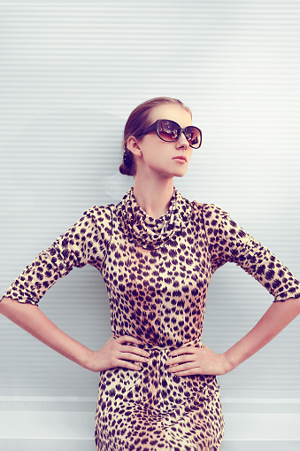 Fashion and people concept - pretty sexy woman in leopard dress and sunglasses posing outdoors in urban style on against the gray wall