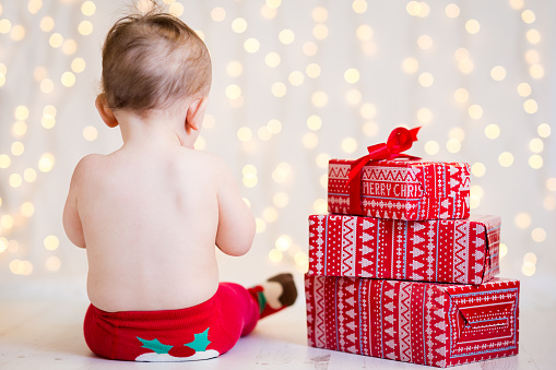 Composition of nicely ribbon wrapped and piled Christmas gift boxes in red and white paper motiv and sitting cute baby from the back in funny festive red tights with knitted Christmas muffin. On white floor with warm and soft bokeh lights as background. Shot on Canon EOS 6D, 85mm prime lens, ISO 400, indoor studio lighting.