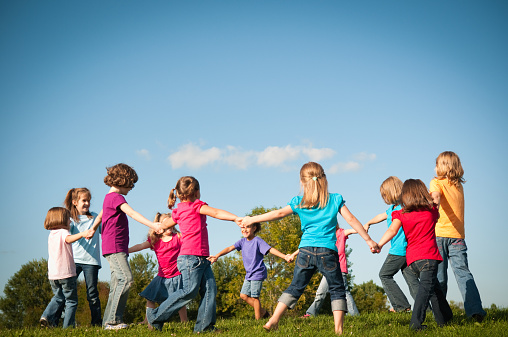 Color photo of a group of eleven happy girls holding hands and spinning in a circle outside on a beautiful summer day with a blue sky in the background.