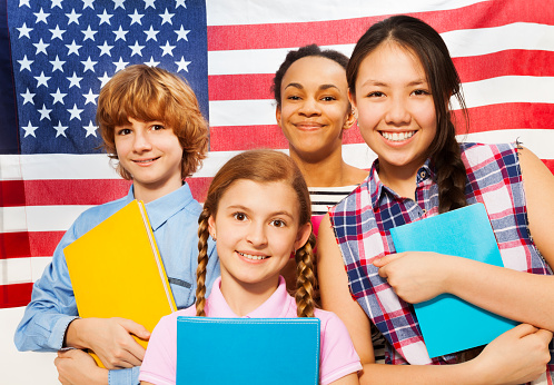 Close-up picture of four smiling multiethnic teenage students standing with textbooks against American flag