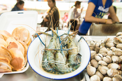 Fresh prawns, and shellfish are set out on the counter of a seafood stand along the traditional floating market in Amphawa,  Thailand. Depth of field is shallow. People are eating in the background.
