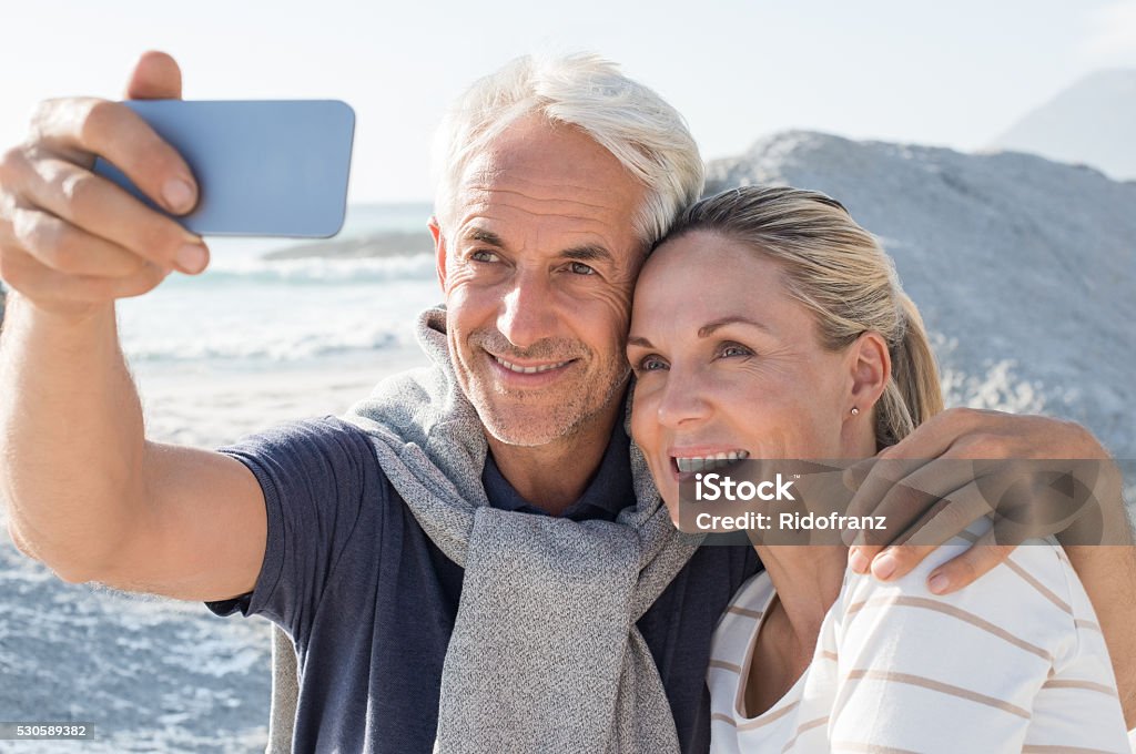 Senior Couple taking selfie Happy romantic couple embracing on the beach and taking a photo with smart phone. Portrait of senior smiling couple taking a selfie in a summer vacation at sea. Happy mature couple take a picture at sunset. Couple - Relationship Stock Photo