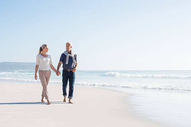 Couple walking at beach Senior couple holding hands at the beach on a bright sunny day. Mature couple in love holding hands and looking each other at the seaside. Smiling wife and happy husband walking barefoot on the white sand. mature couple stock pictures, royalty-free photos & images