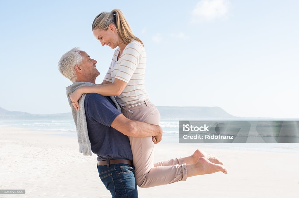 Mature couple in love Cute senior couple hugging on the beach on a sunny day. Happy couple having fun together at the beach. Senior man carrrying her wife at beach and looking at each other. Mature Couple Stock Photo
