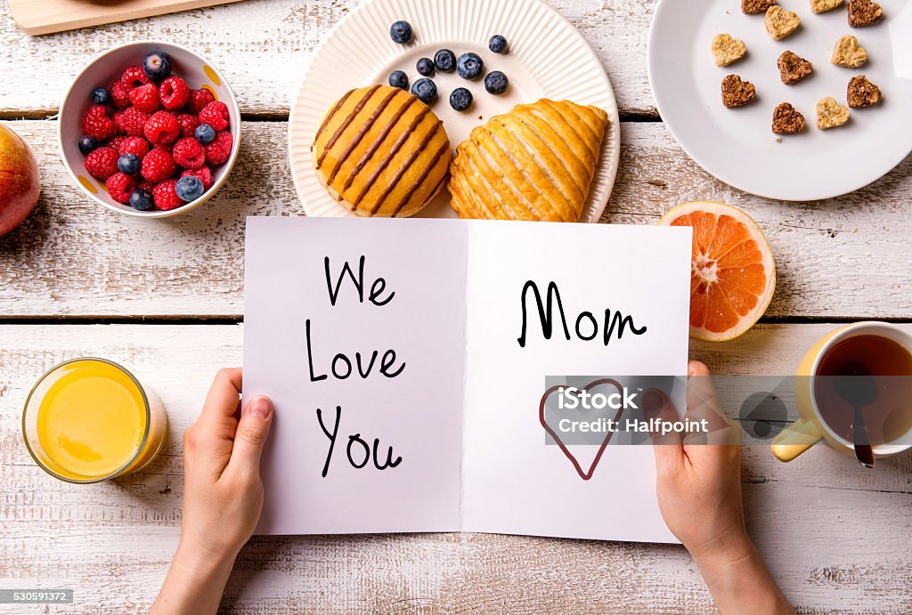 Mothers day composition. Greeting card and breakfast meal. Mothers day composition. Hands of unrecognizable woman holding greeting card with We love you, Mom, text. Breakfast meal. Studio shot on wooden background. Mother's Day Stock Photo