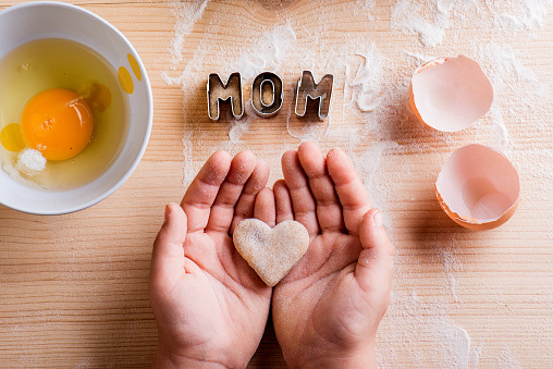 Mothers day composition. Hands of unrecognizable girl baking, holding a heart shaped cookie. Mom sign made of cookie cutters. Egg and flour. Wooden background.