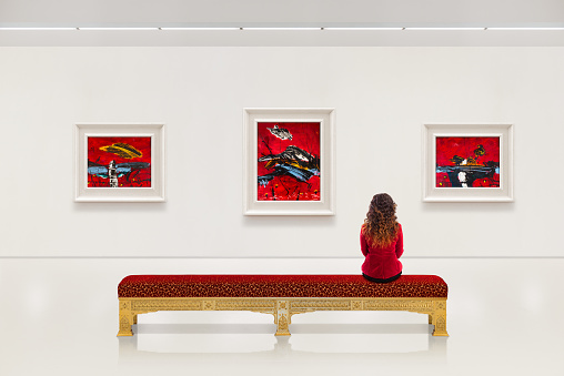In a exhibition centre, lonely young woman wears red jacket, visits an art exhibition and watches artist's collection on the wall. Lightened white wall contains three large frames with red oil fine art paintings.