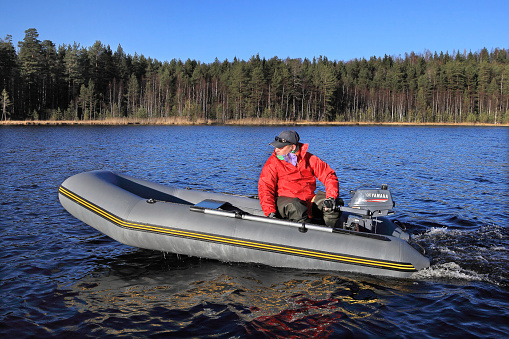 Leningrad Region, Russia - October 20, 2013: One unidentified man in a red cloak, manages gray inflatable rubber boat with an outboard motor on a blue forest lake in the fall on a sunny day.