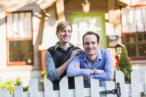 Two gay men standing in the front yard of their home on a bright, sunny day.  They are a happy couple, smiling at the camera.  One man has his arms folded, leaning on the top of a white, picket fence.  His partner is beside him, resting a hand on his shoulder.