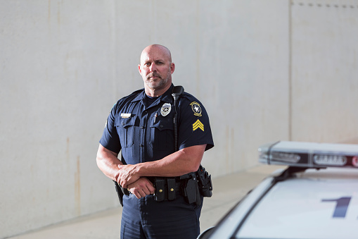 Policeman standing, looking at the camera with a serious expression on his face.  Top of a police car is out of focus in the foreground.  He is in his late forties, with a bald head.