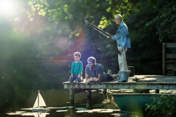 ragazzo che pesca e gioca con la barca a vela giocattolo con padre e nonno al lago - sailing nautical vessel family lake foto e immagini stock
