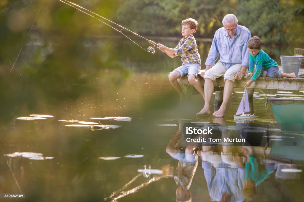 Grandfather and grandsons fishing and playing with toy sailboat at lake  Lake Stock Photo