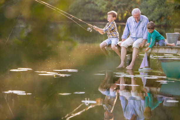 Grandfather and grandsons fishing and playing with toy sailboat at lake stock photo
