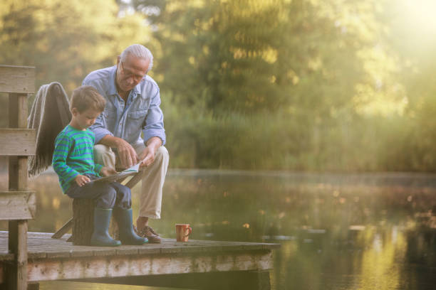 grand-père et petit-fils lisant au lac - leisure activity grandparent grandfather grandson photos et images de collection