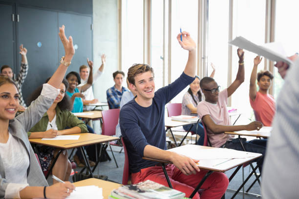 vista de estudiantes sonrientes sentados en escritorios en el aula con los brazos en alto - japanese ethnicity college student asian ethnicity asian and indian ethnicities fotografías e imágenes de stock