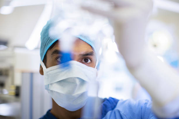 cirujano con máscara quirúrgica, gorra y guantes que observan de cerca el goteo intravenoso - color de ojos fotografías e imágenes de stock