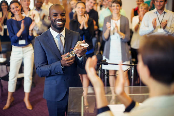 Portrait of young man holding trophy, standing in conference room, smiling to applauding audience  badge photos stock pictures, royalty-free photos & images