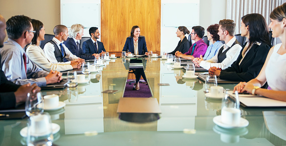 A group of mature adults sit at a boardroom table as they listen in attentively at a corporate business meeting.  They are each dressed professionally and have paper out in front of them as they take notes.