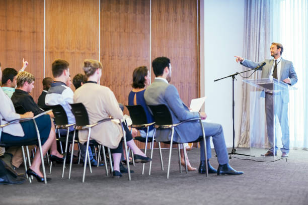 portrait of businessman standing at transparent lectern talking before audience in conference room - board room business conference table window imagens e fotografias de stock