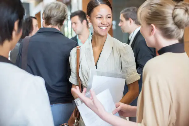 Photo of Portrait of two smiling women holding files and talking, standing in crowded lobby