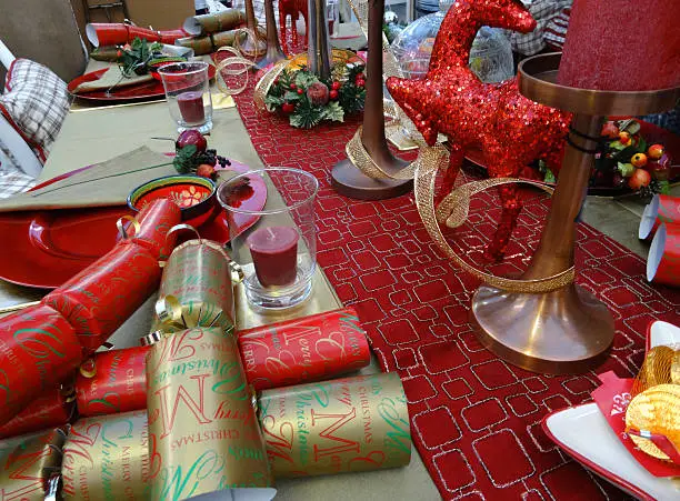 Photo showing a themed dinner table set for a Christmas meal featuring a decorative table cloth / runner, red and gold crackers, red tableware, crockery / plates, paper napkins, wine glasses and red pillar candles, with glittery centrepiece deer ornaments and various seasonal decorations, including golden ribbons.