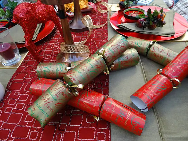 Photo showing a themed dinner table set for a Christmas meal featuring a decorative table cloth / runner, red and gold crackers, red tableware, crockery / plates, wine glasses and red pillar candles, with glittery centrepiece deer ornaments, napkins and plastic fruit.