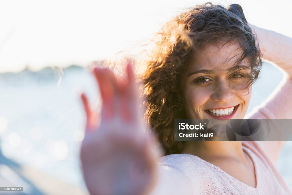 Beautiful young woman standing outdoors by the water Face of a smiling, beautiful young woman with long, dark, curly hair looking at the camera. She is standing outdoors in the sunlight on a dock by the water. It is a windy day, so she has one hand on the back of her head, trying to keep her hair from blowing in her face.  The other hand is extended toward the camera, trying to block the view with her palm out of focus in the foreground. One Woman Only Stock Photo