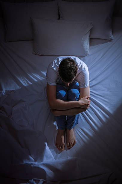 Anxious and scared young man Vertical shot of an anxious and scared young man sitting on his bed at night and contemplating harsh shadows stock pictures, royalty-free photos & images