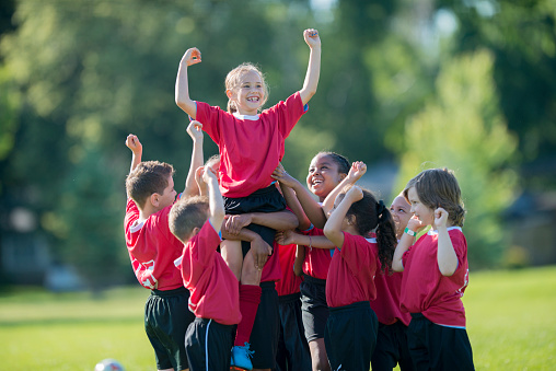 A multi-ethnic group of elementary age children are cheering and lifting a teammate in the air after winning a game.