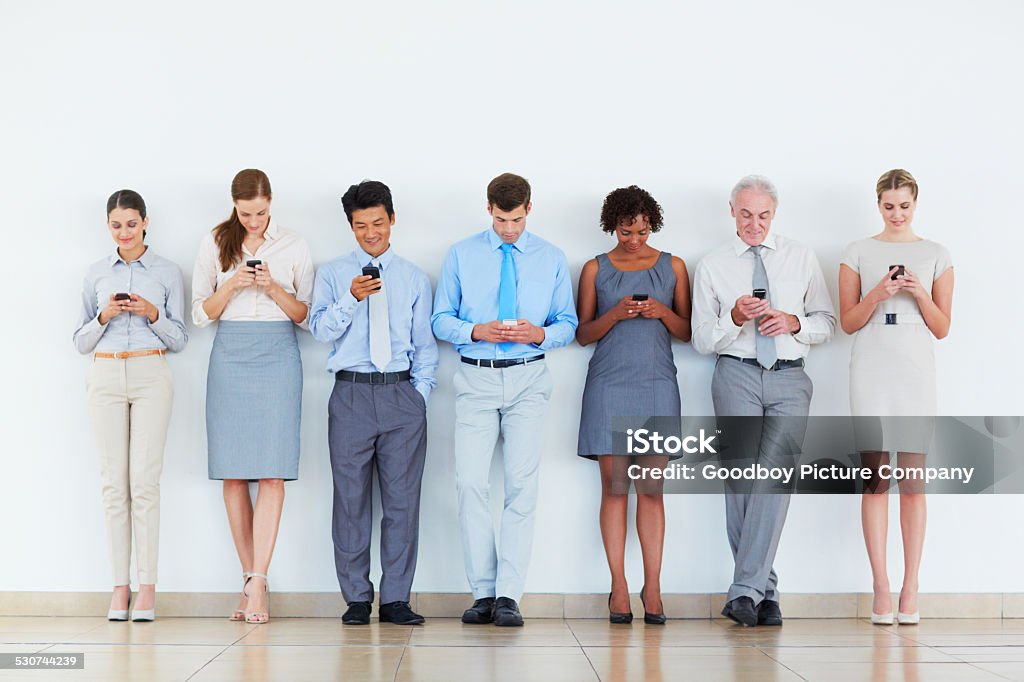 This is what business has become Shot of a group of coworkers all using their mobile devices while leaning against a wall Smart Phone Stock Photo