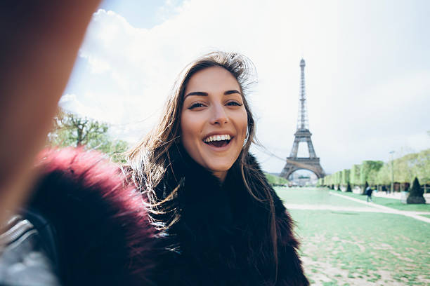 Smiling young woman making selfie in front of Eiffel tower. Hand on camera. With fur coat. Looking at camera. Eiffel tower, park and sunlight on background.