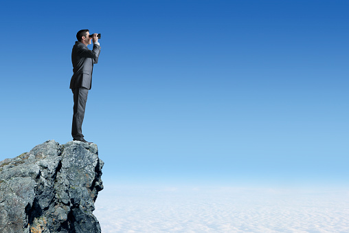 A businessman, standing at a profile to the camera, stands on top of a mountain top high above the clouds as he peers through a pair of binoculars.  The blue sky and clouds provide ample space for copy or text.