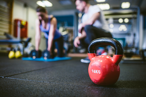 Kettlebell on gym floor. Young woman exercising with personal trainer in back. They are defocused.