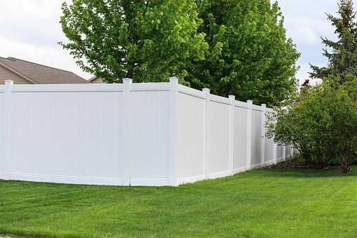 A white vinyl fence running across a yard on spring day with blue sky and trees in the background