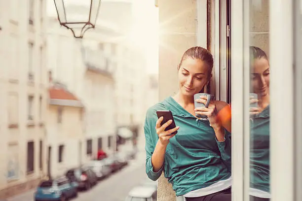 Photo of Happy woman with coffee cup texting on the window