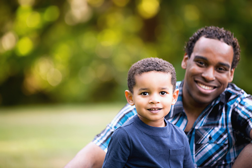 Cute African American father and son smiling in an outdoor park - lots of green background. Looking at camera and happy.