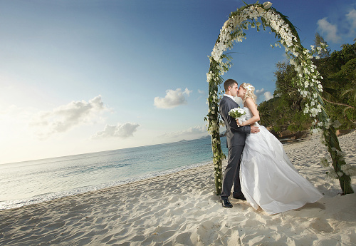 Gorgerous couple kissing, standing under wedding arch during sunset. Beautiful wedding on caribbean island or hawaii.