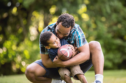 Cute African American father and son smiling in an outdoor park - lots of green background. Looking at camera and happy.Cute African American father and son smiling in an outdoor park - lots of green background. Playing Football
