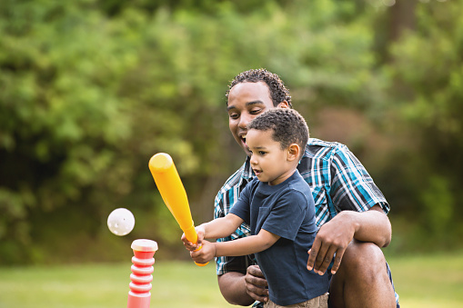 Cute African American father and son smiling in an outdoor park - lots of green background. Playing T Ball.