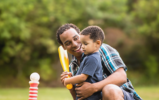 Cute African American father and son smiling in an outdoor park - lots of green background. Playing T Ball.